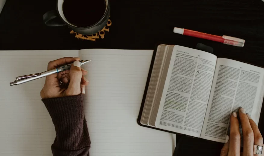 A woman's hand holding an open book and a pen, with a coffee mug placed beside her.