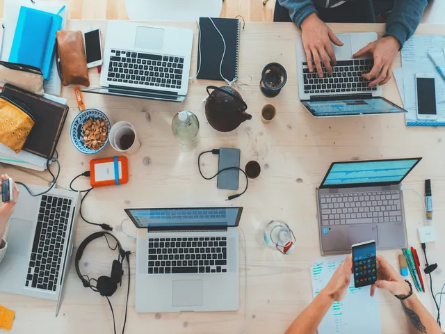 A cluttered workspace with people using laptops and various accessories such as headphones, notebooks, and smartphones on a wooden table