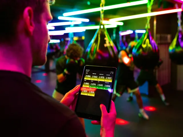 Person-monitoring-data-on-a-tablet-in-a-gym-with-neon-lit-punching-bags-and-people-training-in-the-background